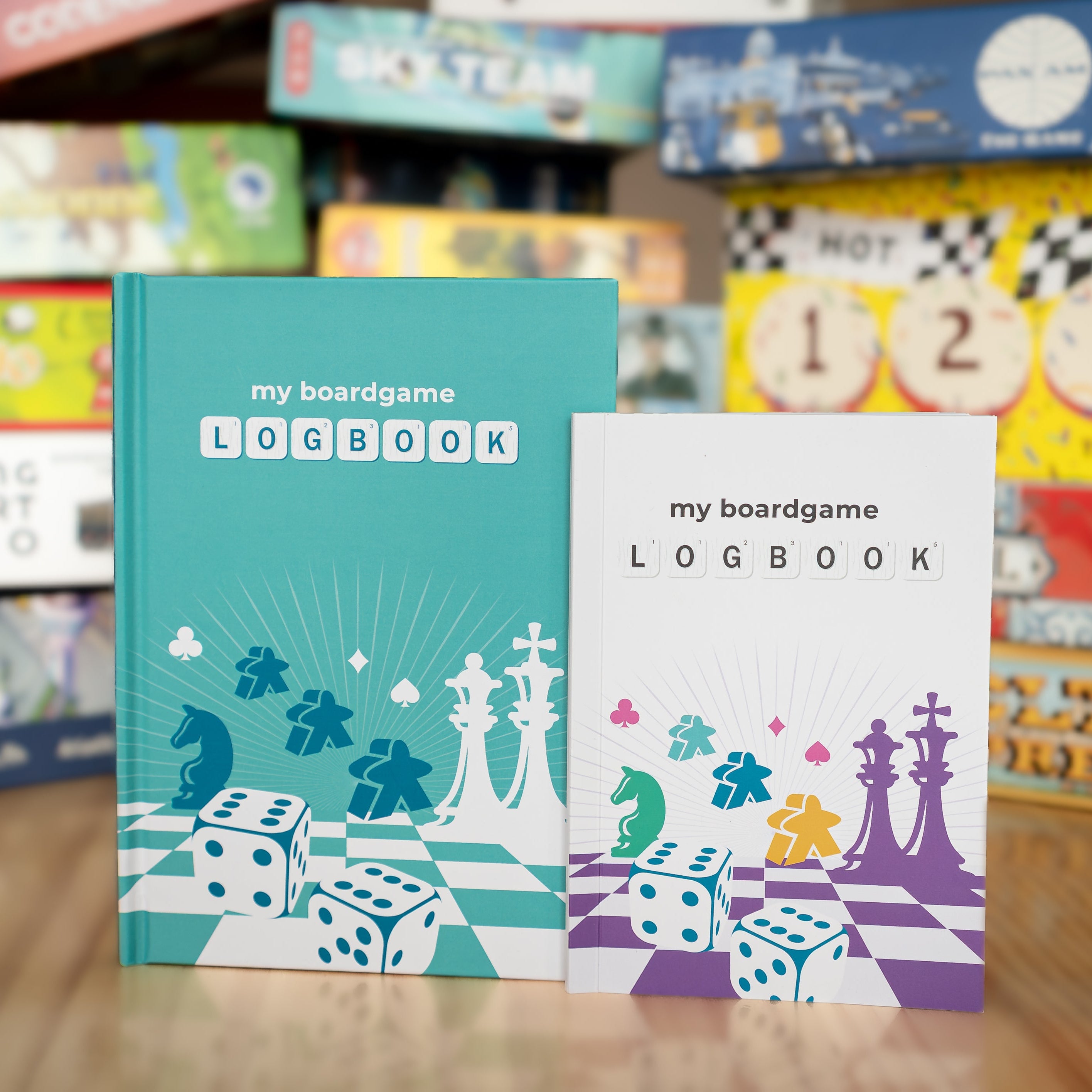 Two 'my boardgame logbook' books with board game-themed designs on a wooden surface with blurred board games in the background.